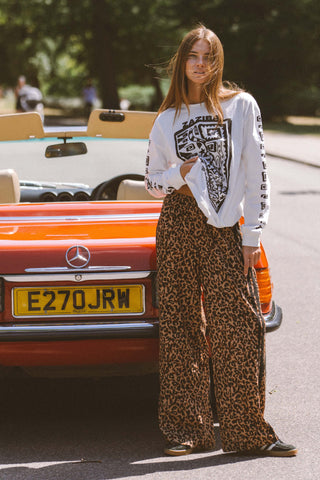 Woman in a white sweatshirt with black graphic design and leopard print pants standing next to a vintage car.