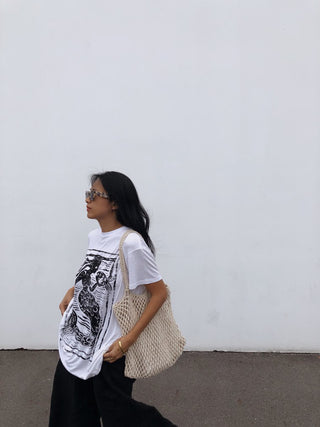 Woman wearing a graphic t-shirt and carrying a beige bag against a white wall.