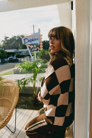 Woman sitting on a wooden deck with a checkered sweater, palm trees, and a 'Dolphins' sign in the background.