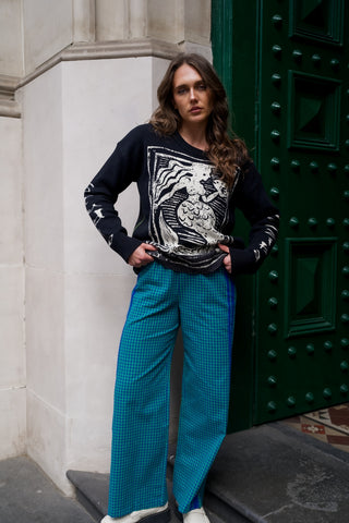 Woman wearing a black sweater with a graphic design and blue pants standing in front of a green door.