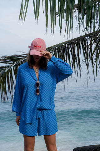 Person wearing a blue checkered outfit and pink cap by the ocean with palm leaves in the background