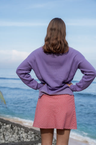 Person wearing a purple long-sleeve shirt and red checkered skirt standing by the ocean.