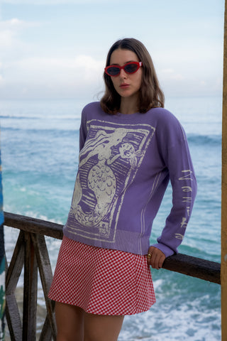Woman wearing a purple sweater with a graphic design and a red checkered skirt, standing by the ocean.