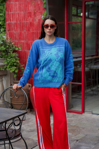 Woman wearing a blue sweater with a dragon design and red pants standing in front of a red building.