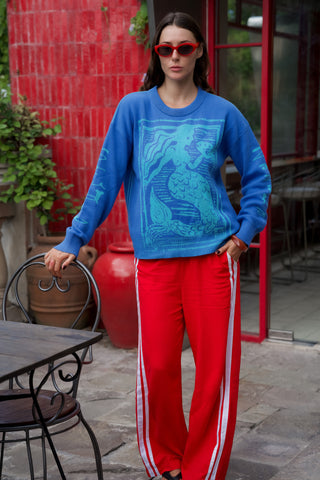 Woman wearing a blue sweater with a graphic design and red pants with white stripes outdoors.