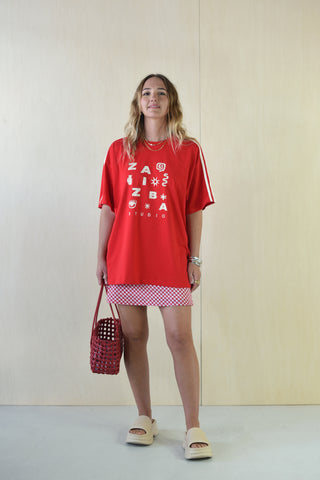 Person wearing a red oversized t-shirt with text and a checkered skirt, holding a red bag against a plain background.