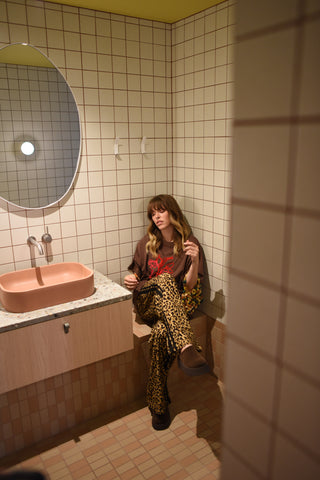 Person sitting on a toilet in a tiled bathroom with a sink and mirror.