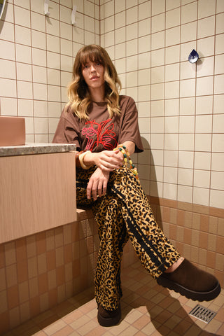 Person sitting on a toilet in a tiled bathroom wearing a brown shirt with a red design and leopard print pants.