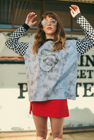 A woman wearing a light blue tie-dye t-shirt with a black snake graphic, a red mini skirt, and a black-and-white checkered long-sleeve shirt underneath, posing outdoors in front of a building wall.