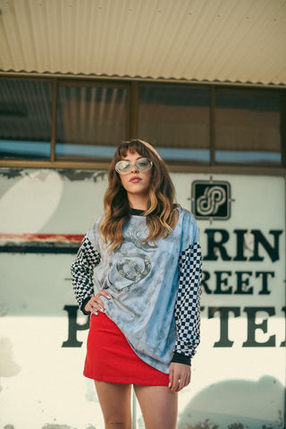 A woman wearing a light blue graphic t-shirt with black-and-white checkered sleeves, a red mini skirt, and round sunglasses, posing in front of a vintage-style building wall.