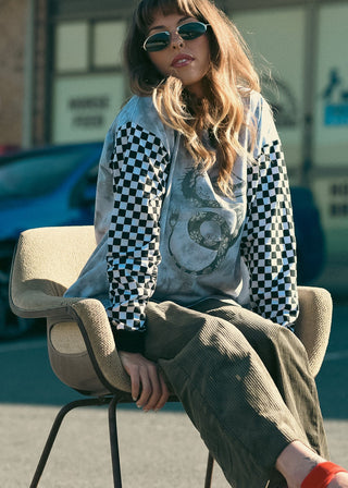 A woman wearing a graphic hoodie with black and white checkered sleeves, a gray graphic t-shirt, and olive green pants sits on a modern beige chair in an outdoor urban setting with cars and storefronts in the background.