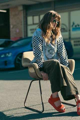 A woman sits on a modern chair outdoors, wearing a light blue and black checkered long-sleeve top, olive green wide-leg pants, and bright red platform sandals. A blue sports car and storefront are blurred in the background.
