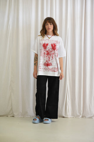 A woman stands in front of a white curtain backdrop wearing an oversized white graphic T-shirt with a red lobster print, loose black pants, and light blue slide sandals.