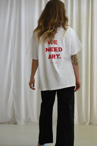 A woman viewed from behind wearing an oversized white T-shirt with bold red text reading 'WE NEED ART.' on the back, paired with black pants, standing in front of a draped white curtain.