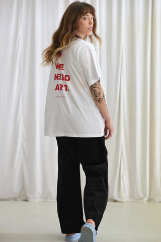 A woman wearing a white oversized t-shirt with red text on the back reading 'WE NEED ART.' and black cargo pants, standing in front of a white curtain backdrop.