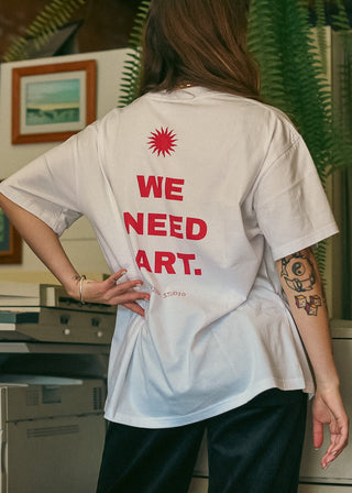 A person viewed from behind wearing an oversized white T-shirt with bold red text on the back reading 'WE NEED ART.' in an office-like interior with printers and framed pictures in the background.
