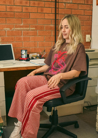 A woman sits in a black office chair in a workspace with a brick wall background, wearing a brown graphic t-shirt, red-and-white gingham pants, and white sneakers. A computer desk, office printer, and framed pictures are visible around her.