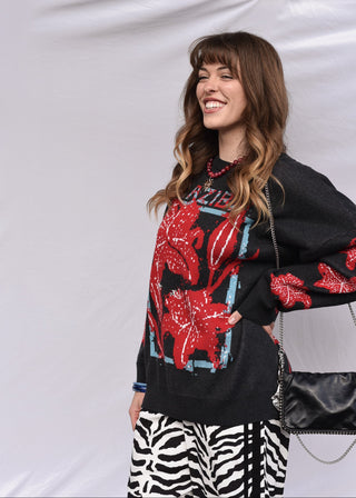 A woman smiling while wearing a black knit sweater with a large red floral pattern and a black-and-white zebra-print skirt, standing against a white fabric backdrop.