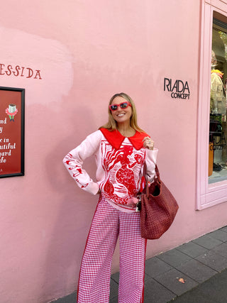 A woman posing outdoors in front of a pink wall, wearing a white and red patterned long-sleeve top, red and white gingham wide-leg pants, and carrying a brown woven handbag.