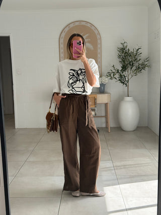 Person wearing a white t-shirt with a graphic design and brown pants in a room.