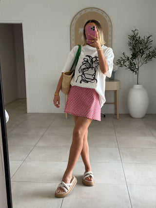 Person taking a mirror selfie wearing a white graphic t-shirt, red checkered skirt, and sandals in a room.