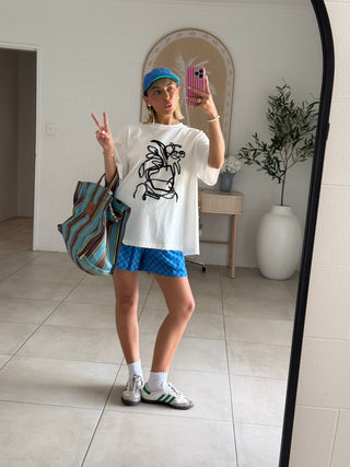Person taking a mirror selfie wearing a white t-shirt with a graphic design, blue shorts, and a blue cap.
