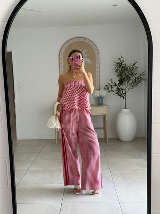 Stylish woman wearing a pink checkered outfit taking a mirror selfie in a room with white walls and a plant.