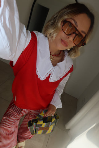 A woman wearing a red knit sweater vest over a white collared shirt, paired with red and white striped trousers, holding a yellow and gray plaid handbag.