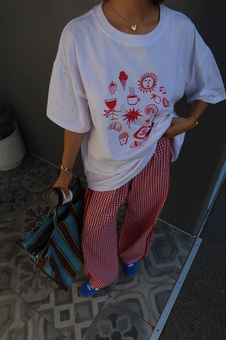 A person wearing an oversized white graphic T-shirt with red illustrations, red-and-white gingham wide-leg pants, and blue shoes, standing on a patterned tile floor next to a dark wall.