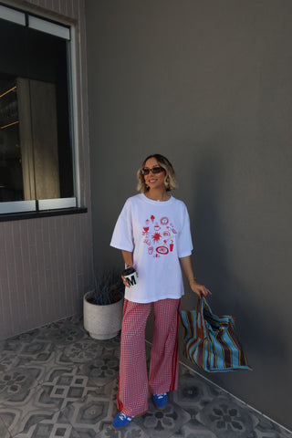 A woman stands on a tiled patio against a gray wall, wearing an oversized white graphic T-shirt with red spider-themed illustrations, red-and-white gingham wide-leg pants, blue sneakers, and holding a striped tote bag.