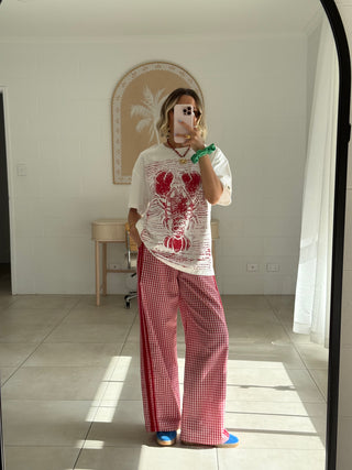 A woman takes a mirror selfie in a bright room, wearing a white graphic t-shirt with a red lobster print and red-and-white gingham wide-leg pants.