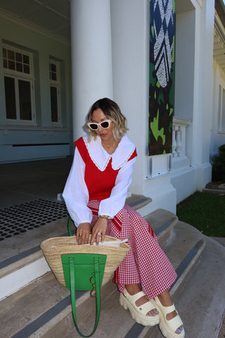 A woman sits on the concrete steps of a white building, wearing a red and white gingham patterned top and matching wide-leg pants, white platform sandals, and white sunglasses. She is holding a green handbag next to a large woven straw tote bag.