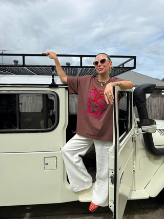 A woman wearing a brown graphic t-shirt, white wide-leg pants, and pink sneakers leans against the open door of a cream-colored vehicle under a cloudy sky.