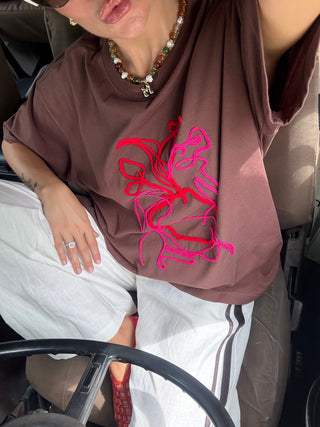 A person sitting in a car wearing an oversized brown T-shirt with abstract pink and red line embroidery on the front, paired with white pants.