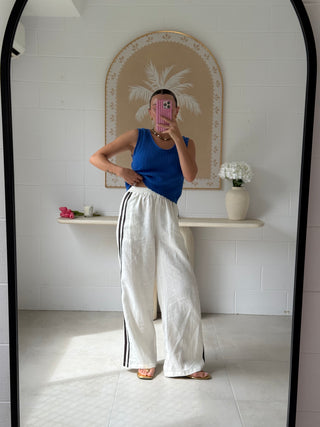 Person taking a mirror selfie wearing a blue top and white pants with black stripes in a room with a decorative wall piece.