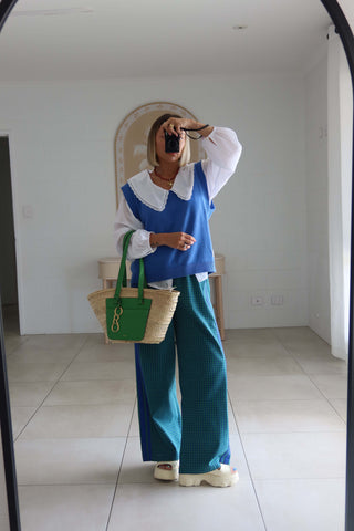 A woman takes a mirror selfie in a minimalist room, wearing a blue and white top with wide teal trousers and holding a green and beige woven tote bag.