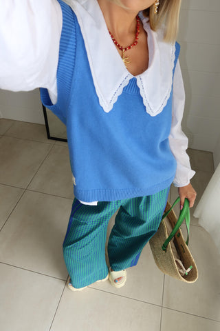 A person wearing a bright blue knit sweater vest over a white long-sleeve shirt with a large white collar, paired with teal corduroy pants and holding a woven straw tote bag with green handles.