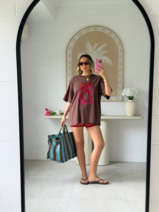 A woman takes a mirror selfie in a bathroom setting, wearing an oversized brown graphic t-shirt with a pink graphic print, red shorts, sunglasses, and holding a striped blue and green tote bag.