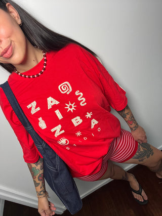 A woman wearing a red graphic t-shirt with white text reading 'ZAZIBA STUDIO' and matching red-and-white striped shorts, styled with a dark denim jacket and black sandals.