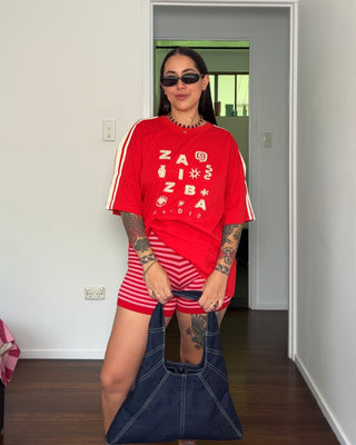 A woman stands indoors wearing a bright red oversized graphic T-shirt with white lettering and red-and-white striped shorts, holding a large navy tote bag. She is in a doorway with white walls and dark wood flooring visible.