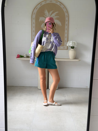 Person taking a mirror selfie wearing a purple sweater, green shorts, and a pink hat in a bathroom.