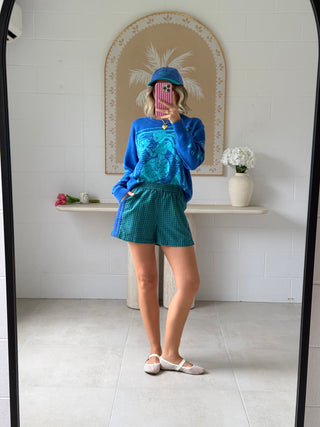 Person taking a mirror selfie wearing a blue sweater and green shorts in a room with a white tiled wall and decorative elements.