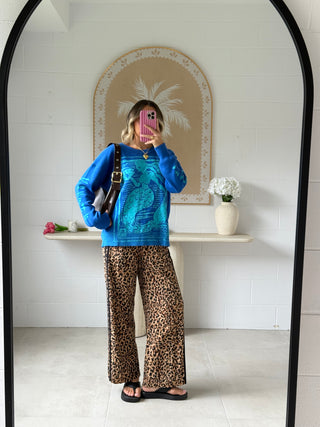 Person wearing a blue sweater with a graphic design and leopard print pants, standing in front of a mirror.