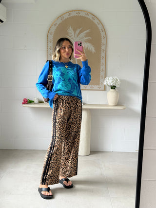 Woman taking a mirror selfie wearing a blue top and leopard print pants in a room with a decorative mirror and shelf.