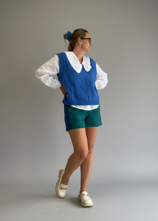 Woman wearing a blue vest over a white blouse with green shorts against a plain background