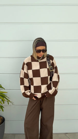 Person wearing a brown and beige checkered sweater with a belt, standing against a light gray wall.