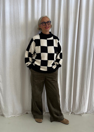 A person wearing a black and white checkered knit sweater, olive green wide-leg pants, and brown shoes stands against a white draped curtain backdrop.