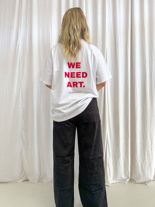A person standing with their back to the camera, wearing an oversized white t-shirt with bold red text reading 'WE NEED ART.' on the back, paired with loose black pants, set against a backdrop of white draped curtains.