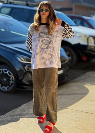 A woman stands outdoors in a parking lot wearing a long-sleeve graphic top with a black and white checkered pattern on the sleeves and a faded light gray body, paired with wide-leg brown pants and red slide sandals.