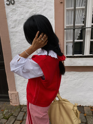 Person wearing a red vest over a white shirt with ruffled details, standing in front of a building.